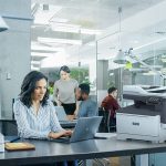 Xerox® C325 Farb-Multifunktionsdrucker in einer Bürosituation Berufstätige Frau tippt auf einem Laptop am Schreibtisch in einer Büroumgebung neben einem Xerox® C325-Drucker